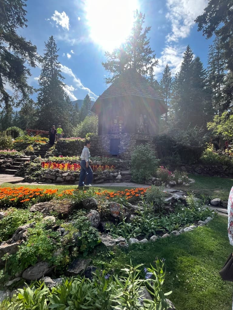 A sunlit botanical garden in Banff with flower beds, a wooden gazebo, evergreen trees, and a mountain peak.