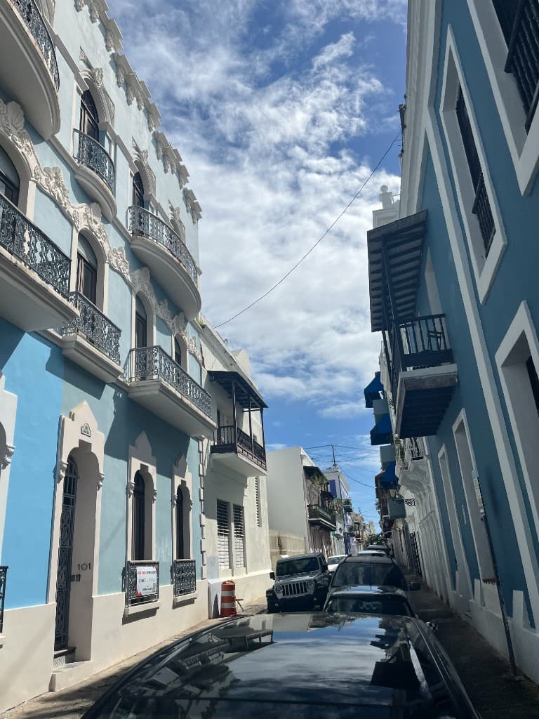 A narrow sunlit street in Old San Juan lined with blue colonial buildings, white trim, and wrought-iron balconies.